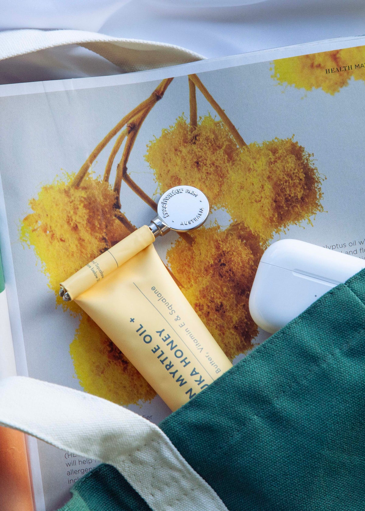 A styled flat-lay of a cream-colored canvas tote bag containing a green fabric pouch, a yellow tube of body oil, a white earbud case, and a printed magazine, all arranged on white fabric.